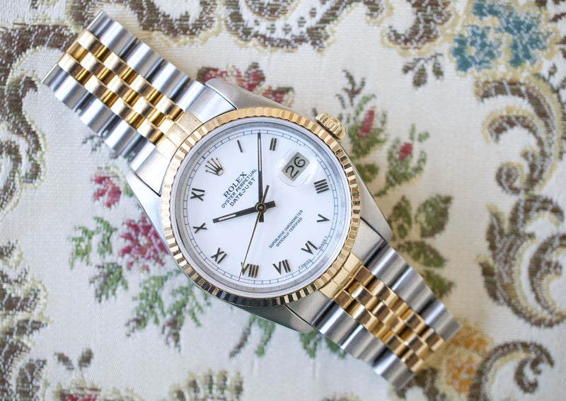Two-tone luxury watch with Roman numerals and date display on a floral background