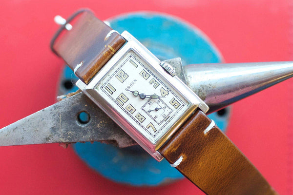 Vintage rectangular Gruen watch with a brown leather strap on a blue and red background.
