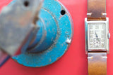 Vintage rectangular Gruen watch with brown leather strap on a red background.