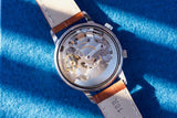 Rear view of a luxury Swiss-made watch movement with a tan leather strap on blue fabric.