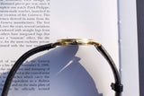 Side view of a gold luxury watch with a black leather strap resting on a book page.