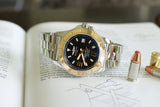 Stainless steel luxury watch with black dial and gold bezel on open book, next to bullets