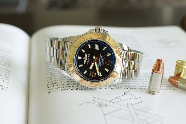 Stainless steel luxury watch with black dial and gold bezel on open book, next to bullets