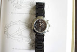 Black chronograph luxury watch with metal bezel and rubber strap on a technical drawing