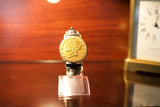 Vintage gold coin watch with a female profile, dated 1904, on a clear display stand.