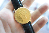 Gold coin luxury watch with 1904 date and black leather strap held in a hand