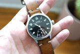 Close-up of a vintage Heuer watch with a black dial and brown leather strap on a wrist.
