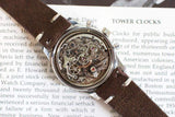 Vintage chronograph watch with visible mechanical movement and brown suede strap on an open book