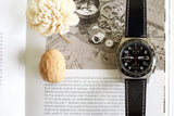 Luxury chronograph watch with black leather strap on open book next to a flower and nut