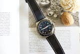 Luxury chronograph watch with black leather strap on open book, flower and nut beside