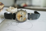 Vintage Swiss chronograph watch with patina dial and black leather strap on a sketchbook
