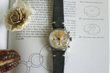 Vintage chronograph watch with leather strap on technical book, floral accent nearby