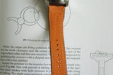 Brown leather watch strap with stitching and embossing, shown over watchmaking manual