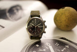 Luxury pilot chronograph watch with black dial and leather strap, displayed on an open book.