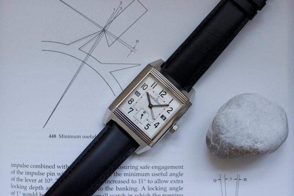 Rectangular silver luxury GMT watch with black leather strap on technical drawing and book