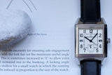 Rectangular luxury watch with a black leather strap on an open book, silver case, white dial.