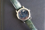 Luxury silver watch with black dial and green leather strap on dark leather surface