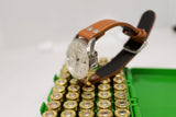 Vintage chronograph watch with brown leather strap displayed on gold ammunition rounds in green box