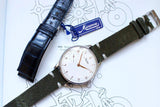 Minerva 140th Anniversary limited edition men's watch with white dial, olive suede strap, next to black leather strap and blue Minerva tag
