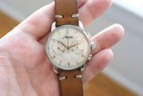 Close-up of a vintage Minerva chronograph watch with a brown leather strap held in a hand.
