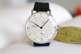 Minimalist luxury watch with white dial, small seconds subdial, date display, and black leather strap