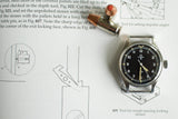 vintage Omega Fat Arrow military watch with black dial on technical manual page, bullet nearby