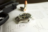 vintage military-style watch with black dial on technical manual, nearby bullet and metal object