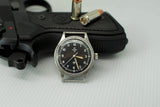 Omega Fat Arrow black dial military watch with bullets and handgun on white surface