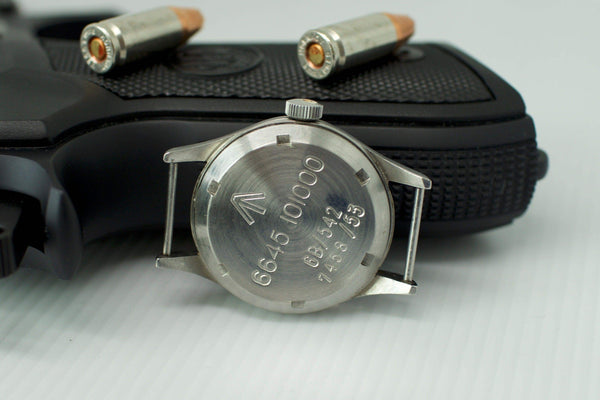 Back view of vintage Omega Fat Arrow watch case with engraved markings, placed on a handgun with bullets