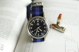 Omega vintage military watch with blue NATO strap on open book, next to two bullets
