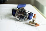 Omega Fat Arrow vintage military watch with black dial and blue NATO strap on an open book beside two bullets