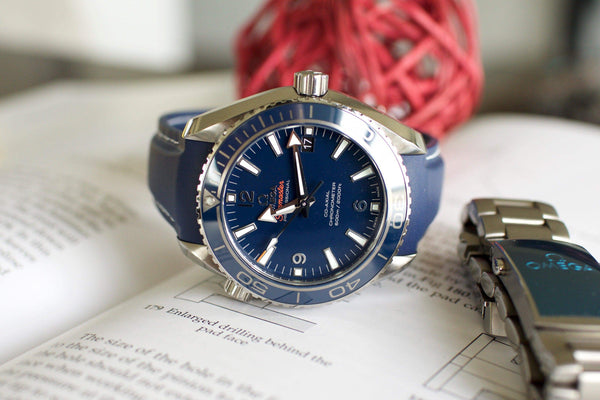 Blue Omega Seamaster luxury dive watch with rubber strap on open book, metal bracelet beside