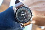 Close-up of an Omega Speedmaster Professional luxury watch with a brown leather strap on a wrist.