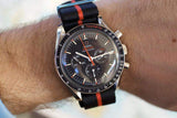 Luxury Swiss chronograph watch with black dial and orange accents on striped nylon strap, worn on wrist