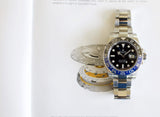 Stainless steel GMT watch with black and blue bezel on open book, mechanical parts visible.