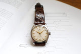 Vintage luxury wristwatch with cream dial and brown leather strap on technical drawing.