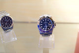 Two luxury Rolex watches displayed on transparent stands against a light background.