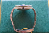 Side view of a luxury two-tone watch with a blue bezel on a green textured background.