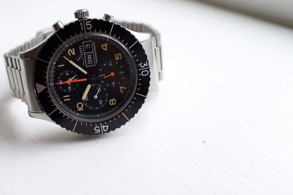 Sinn 156 pilot chronograph watch with black dial, tritium markers, and steel bracelet on white surface