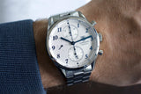 Silver chronograph luxury watch with white dial and blue numerals on wrist