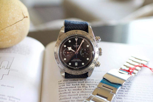 Luxury chronograph watch with black dial and fabric strap displayed on an open book