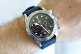 Stainless steel chronograph watch with black dial on blue strap, worn on wrist.