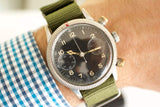 Vintage pilot watch with black dial, chronograph subdials, and green fabric strap on wrist