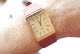 Close-up of a gold Rolex Cellini square watch with a red textured leather strap on a wrist.