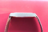 Side view of a luxury watch with a rectangular silver case and brown leather strap