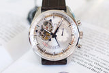 Silver chronograph luxury watch with visible movement, brown leather strap, on open book