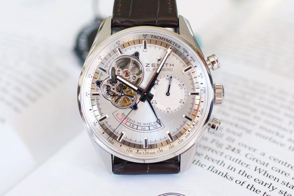 Silver chronograph luxury watch with visible movement, brown leather strap, on open book